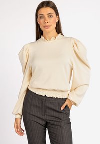 Crème-kleurige pullover met pofmouwen en een gerimpelde halslijn. Heeft een gerimpelde zoom en is gecombineerd met grijzige broek met fijne streep.