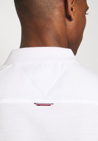 Bílá polo košile s límečkem, která má na zadní straně trojúhelníkový design a malou červenou, bílou a modrou insignii blízko výstřihu.