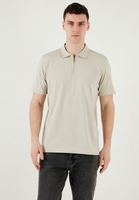Beige Polo-Shirt mit kurzen Ärmeln und Reißverschlusskragen, glatter Textur und tailliertem Schnitt, kombiniert mit dunklen Jeans.
