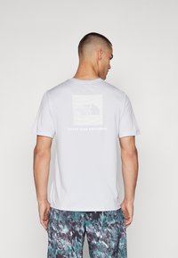 Heled halli lühikeste varrukatega t-särk, mille taga on ruudukujuline logo trükk ja selle all tekst "ÄRA PEATU UURIMISEL".