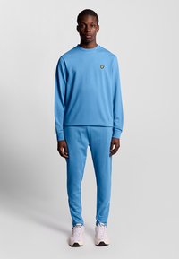 Blauwe sweatshirt en bijpassende broek van zacht materiaal, met een klein gele vlinderlogo op de borst. Sneakers zijn wit met subtiele accenten.