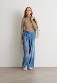 Un pull à col roulé en tricot beige, un pantalon en denim bleu clair à jambes larges et des baskets grises. Le modèle tient une veste légère, debout contre un fond uni.