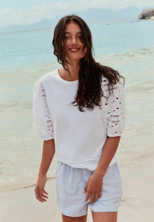 Kobieta z długimi ciemnymi włosami, ubrana w biały top z koronkowymi rękawami i jasne, w paski szorty, stoi uśmiechnięta na piaszczystej plaży z spokojnym morzem w tle.