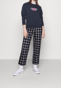 Navy crewneck sweatshirt with "TOMMY JEANS" logo, paired with black and white checkered wide-leg pants and white high-top sneakers.