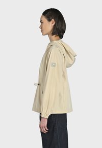 Giacca leggera beige con cappuccio, vita regolabile con cordino, polsini elasticizzati e una patch con logo sulla manica sinistra. Tessuto liscio, design casual.