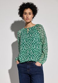 Grüne gemusterte Bluse mit einem Schlüssel kreuz-Ausschnitt und langen Ärmeln, aus leichtem Stoff. Kombiniert mit dunkelblauen Jeans.