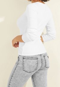 Donna che indossa una camicia bianca attillata a maniche lunghe e jeans a vita alta grigi chiari, posizionata di lato su uno sfondo semplice.