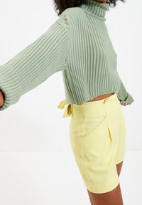 Maglione verde a coste con collo alto e maniche larghe, abbinato a una mini gonna giallo chiaro con lacci laterali e pieghe nette.