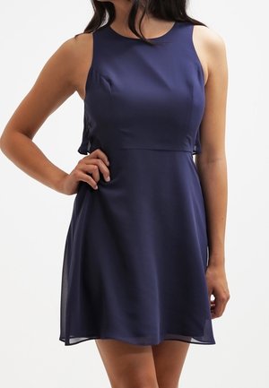 Mouwloze, marineblauwe jurk tot op de knieën met een aansluitend lijfje en een licht uitlopende rok, gedragen door een persoon met de hand op de heup.