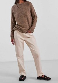 En person står klädd i en brun tröja, krämfärgade byxor och svarta sandaler, poserad i en neutral miljö med en ljus bakgrund.