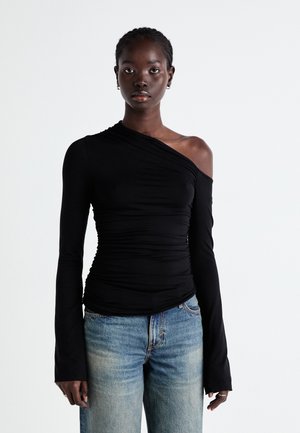 Haut asymétrique noir à manches longues en tissu doux avec des détails froncés sur une épaule, porté avec un jean bleu.