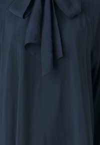 Tummansininen pusero, jossa on pehmeä kangas ja voimakas etuosan rusettidetaili. Tekstuurilla näyttää olevan sileä ja virtaava ilme.