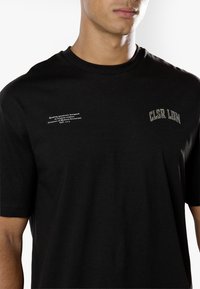 Άνδρας που φοράει μαύρο t-shirt με μικρό λευκό κείμενο στο αριστερό στήθος και γκρι κείμενο "CLSR LDN" στο δεξί στήθος.