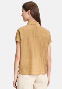 Camicia a maniche corte con colletto in un tessuto marrone chiaro stropicciato. La vista posteriore mostra dettagli di pieghe e una vestibilità rilassata, abbinata a pantaloni chiari.