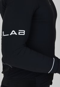 Zwart lange mouwen sporttop gemaakt van rekbare stof. Heeft een contrasterend wit logo op de arm en een subtiele witte accent op de pols.