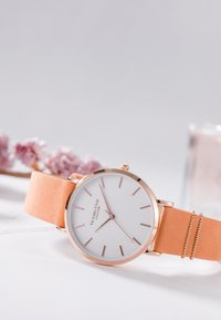 Roséguld klocka med en vit urtavla, smala timmarkörer och ett persikofärgat suèdearmband med en minimalistisk design och spänne.