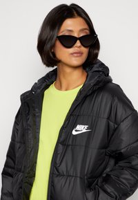 Svart pufferjakke med hette og hvit Nike-logo, båret over en lys limegrønn skjorte. Sorte katteøye-solbriller fullfører stilen.
