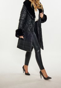 Czarna pikowana kurtka puchowa z kołnierzem i mankietami z sztucznego futra; zestawiona z dopasowanymi czarnymi legginsami i butami na obcasie w groszki. Miękka faktura, długi fason.