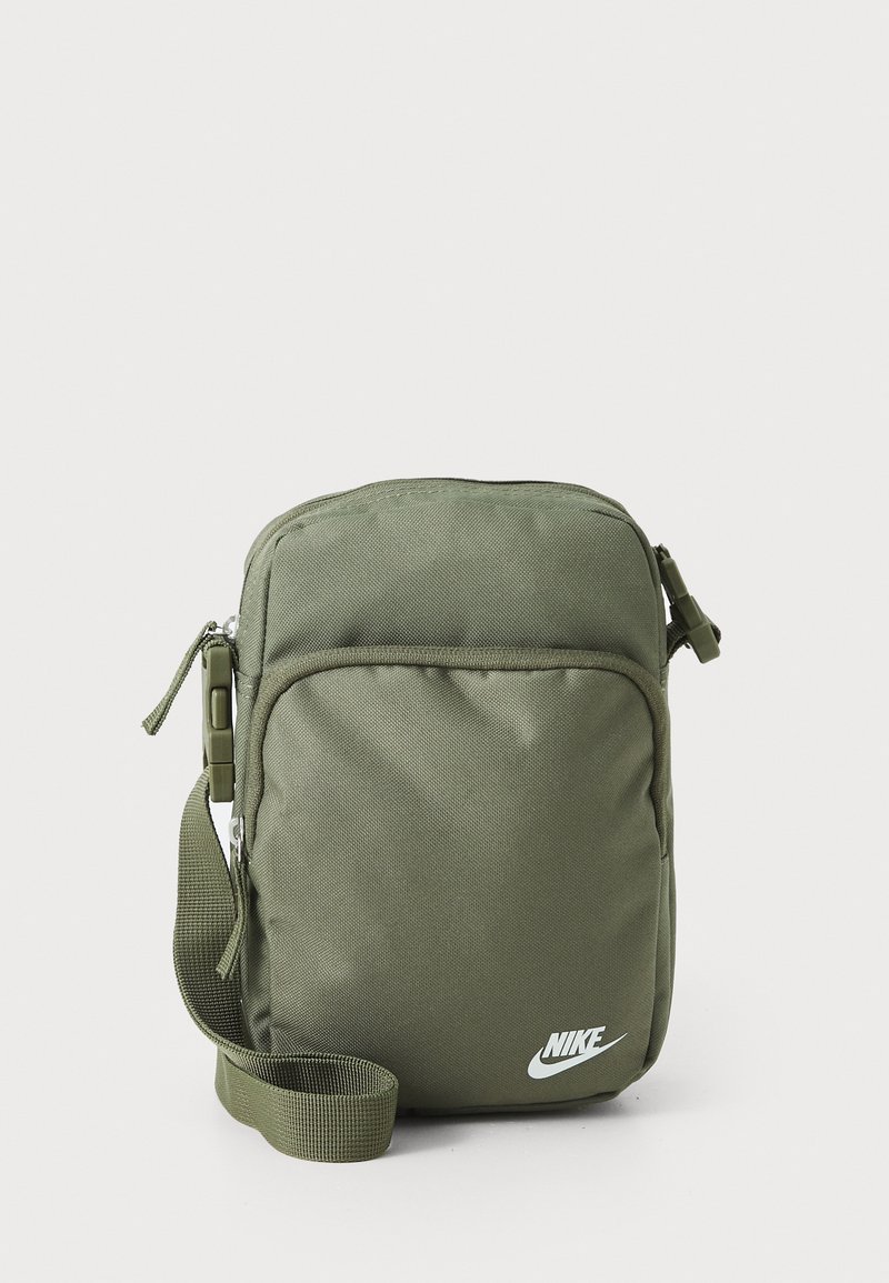 Zaļš auduma plecu soma ar regulējamu siksnu, rāvējslēdzēja aizdare, priekšējais kabata un balts Nike logo apakšā priekšpusē.