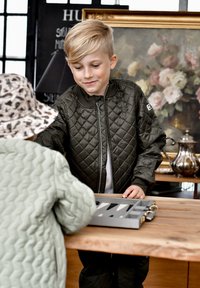 Deti sa venujú činnosti pri drevenom stole. Jeden má na sebe prešívanú olivovo zelenú bomber bundu; druhý textúrovaný svetlozelený kabát a vzorovanú čiapku.