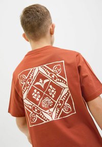 Rūsu krāsas T-krekls ar lielu baltu grafisko printu uz muguras, kas satur ziedu un ģeometriskus dizainus, īsām piedurknēm un apaļu kaklu.