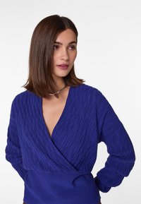 Blaue, gestrickte Wickeljacke mit strukturiertem, geripptem Muster, tiefem V-Ausschnitt und langen, puffigen Ärmeln, getragen mit einer schlichten Silberkette.