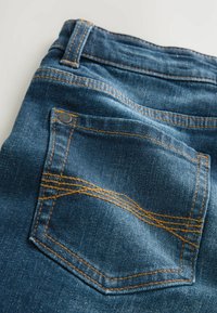 Jean en denim bleu foncé avec des coutures orange, présentant une poche arrière avec une couture épaisse et angulaire. Tissu texturé avec un léger éclat.