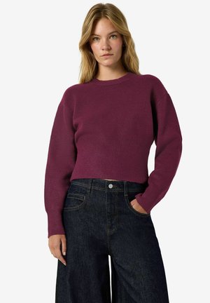 Femme aux cheveux clairs portant un pull en maille violet à manches longues et un jean taille haute bleu foncé, debout avec une main dans la poche.
