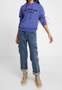 Felpa viola con la scritta "American Eagle", abbinata a jeans cargo blu con tasche laterali e stivali beige con lacci.
