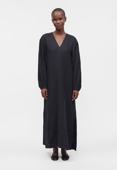 Femme debout portant une longue robe noire à manches longues, un col en V, une fente latérale et des chaussures plates noires sur un fond clair uni.