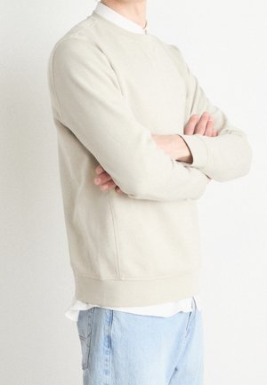 Persona con le braccia incrociate che indossa una felpa beige sopra una camicia bianca e jeans azzurri, in piedi davanti a uno sfondo bianco.