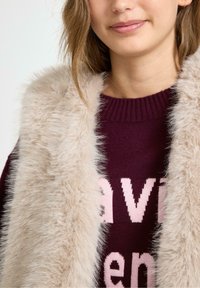 Fluffy beige nepbont vest over een kastanjebruine trui met roze letters, met een geribbelde halslijn en contrasterende texturen.