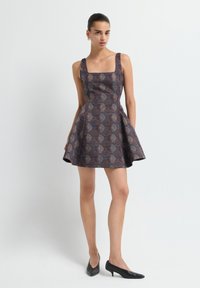 Robe avec un décolleté carré, corsage ajusté et jupe évasée dans un motif sombre, présentant des nuances de bleu et de marron. Portée avec des talons noirs.
