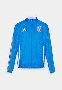 Blå vindjacka med vita detaljer, hela dragkedjan, Adidas-logotyp, emblem för det italienska landslaget och flerfärgade ränder på ärmarna.