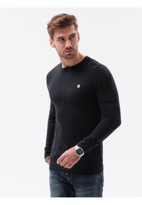 Sort langærmet T-shirt lavet af bomuld, med rund halsudskæring og lille hvidt logo på brystet, parret med blå jeans og et ur.
