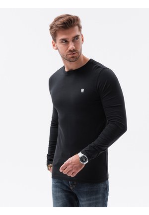 Schwarzes langärmeliges T-Shirt aus Baumwolle, mit rundem Halsausschnitt und kleinem weißen Logo auf der Brust, kombiniert mit blauen Jeans und einer Uhr.