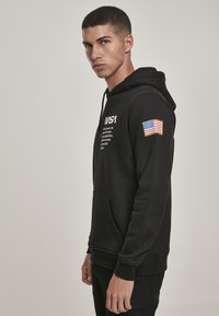 Sweat-shirt à capuche noir avec poche avant, logo NASA en blanc et patch du drapeau américain sur la manche droite. Texture de tissu lisse.