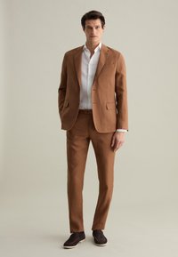 Completo in lino marrone con giacca su misura e pantaloni slim-fit, abbinato a una camicia bianca. Le scarpe in camoscio senza lacci completano l'outfit.