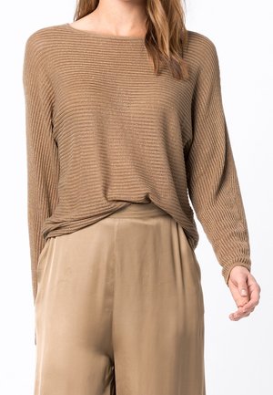 Femme portant un haut côtelé à manches longues de couleur marron clair, rentré dans un pantalon beige lisse taille haute, sur fond blanc.