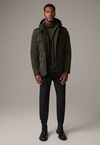 Olivgrüne Pufferjacke mit Kapuze und mehreren Fronttaschen, getragen über einem dunkelgrünen Pullover, kombiniert mit schwarzen schmal geschnittenen Hosen und schwarzen Stiefeln.