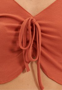 Top color ruggine con texture a costine, caratterizzato da una scollatura a V profonda e un dettaglio a nodo al centro, accentuato da bordi smerlati.