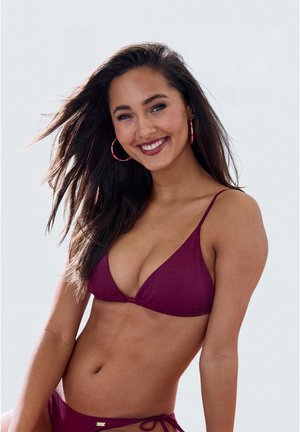 Mujer sonriente con cabello largo y oscuro lleva un bikini color granate y pendientes de aro rojos, posando contra un fondo claro liso.