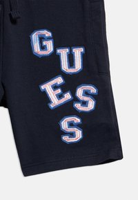 Σκούρο μπλε ύφασμα με μεγάλους ραμμένους λευκούς χαρακτήρες περιγραμμμένους με μπλε και κόκκινο, σχηματίζοντας τη λέξη "GUESS" τοποθετημένη διαγώνια.