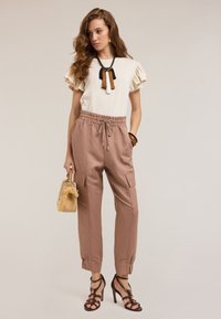 Beige topp med volanger på ärmarna, mjuka bruna vävda byxor med snörning, remsandaler och en liten fransad väska med ett trähandtag.