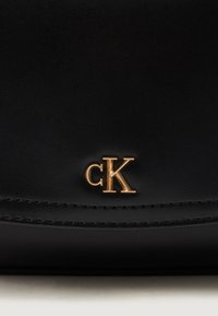 Borsa a spalla in pelle nera con logo dorato "CK", texture liscia e dettagli di cucitura discreti lungo il bordo superiore.