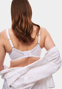 Reggiseno bianco in pizzo con spalline regolabili, caratterizzato da un motivo floreale e chiusura a gancio. Il modello è parzialmente coperto da una camicia bianca.