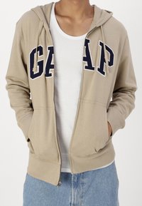 Sweat à capuche beige zippé avec le logo "GAP" en bleu marine, fabriqué en tissu mélangé de coton. Comprend une capuche à cordon de serrage et des poches avant.