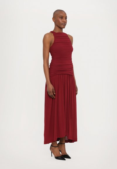 Burgunderfarbenes ärmelloses Kleid mit gerafftem Oberteil und fließendem Rock. Weicher Stoff, knöchellang, getragen mit schwarzen spitz zulaufenden High Heels.