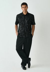 Schwarzes Kurzarmshirt mit Pailletten, figurbetont, kombiniert mit schwarzen weit geschnittenen Hosen und glänzenden schwarzen Schuhen. Heller Hintergrund.