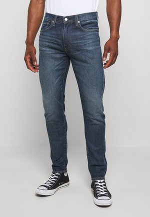 Homme portant un jean slim bleu foncé, un t-shirt blanc et des baskets montantes noires sur un fond clair uni.
